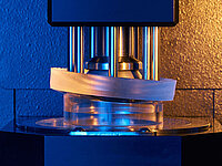 Machine pressing or shaping a glass lens in a precise manufacturing process.