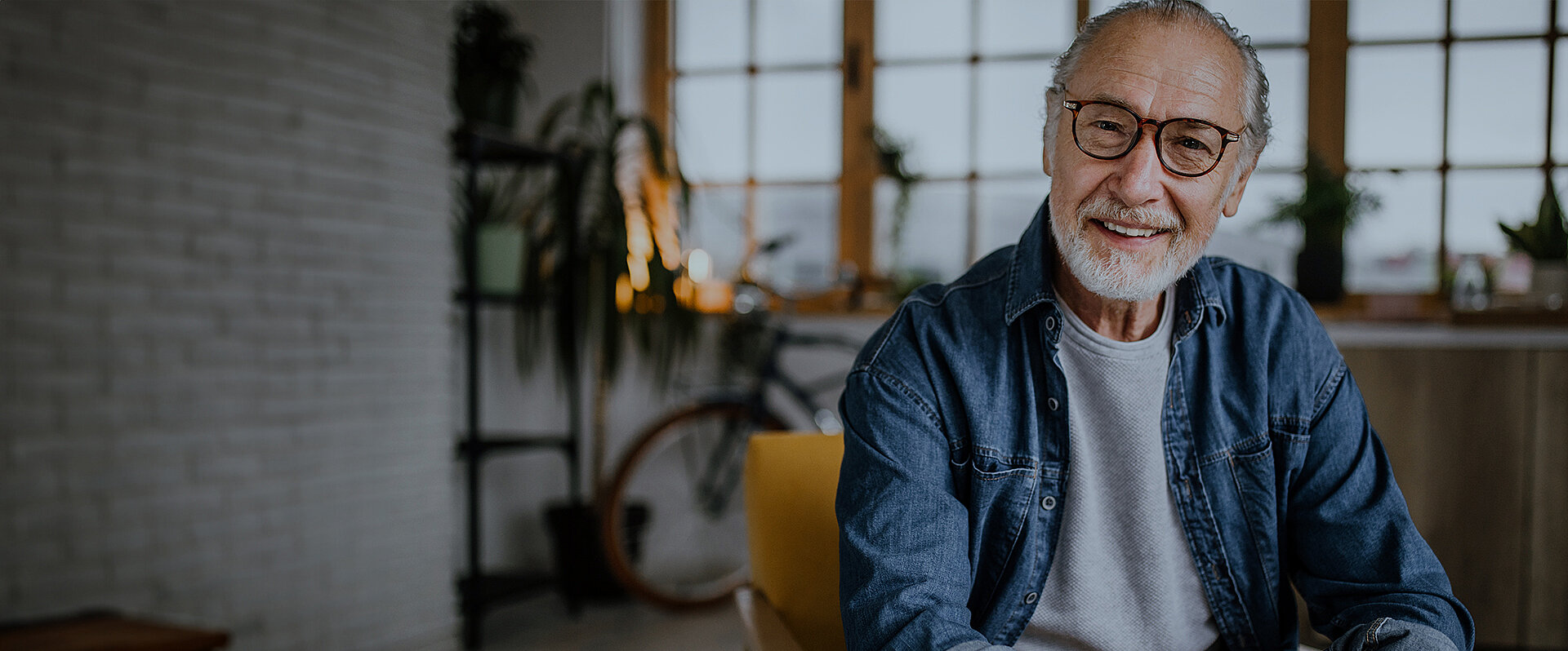 Smiling elderly man with glasses and a beard, sitting indoors in a bright room with plants and a bicycle in the background.