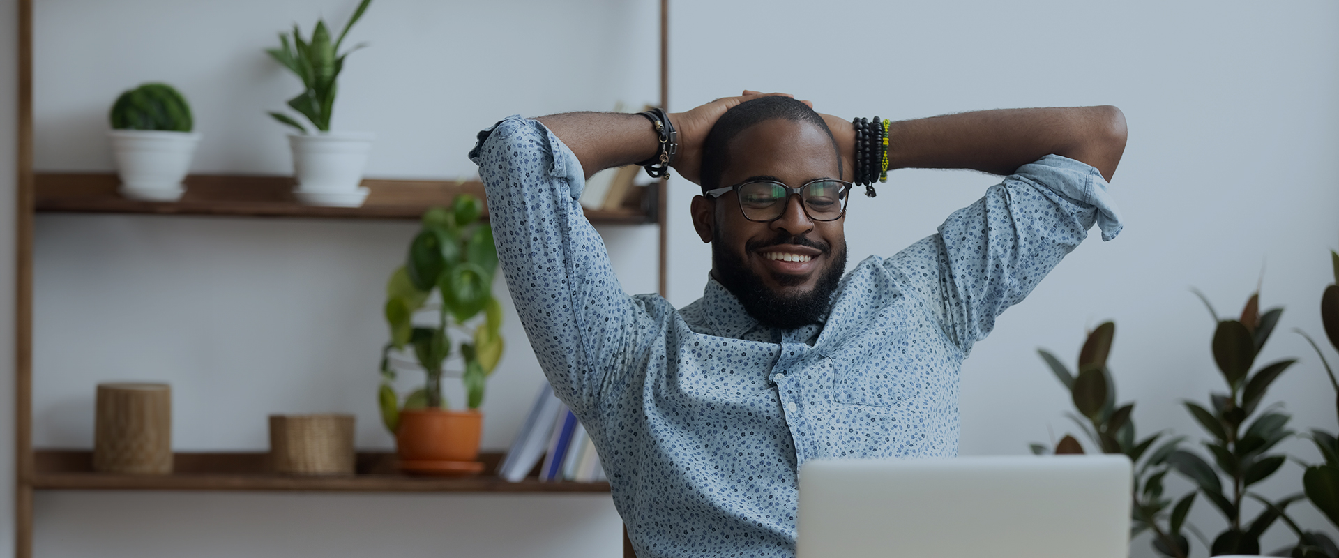 Smiling man with glasses sitting at a desk, stretching with his hands behind his head, with plants on shelves in the background